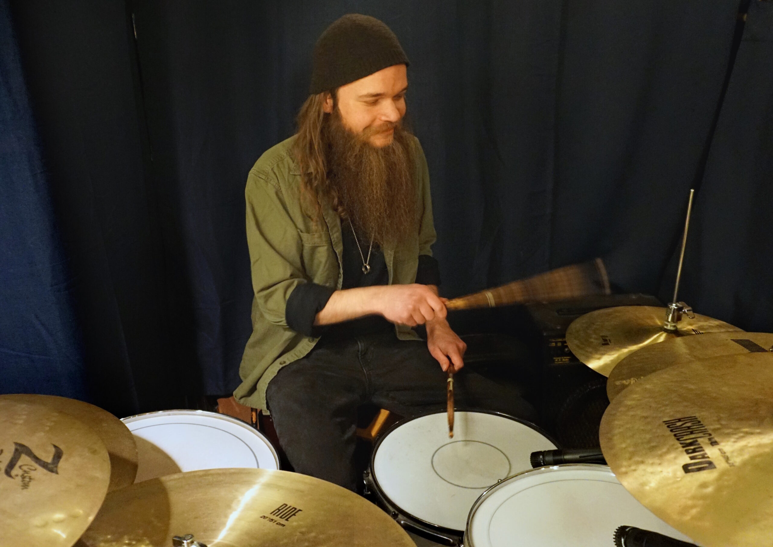 Cassidy Jay sits at a kit, drumsticks in each hand. His right hand is blurred as it hits a cymbal. He has a smile on his face, with his head turned away from the camera.